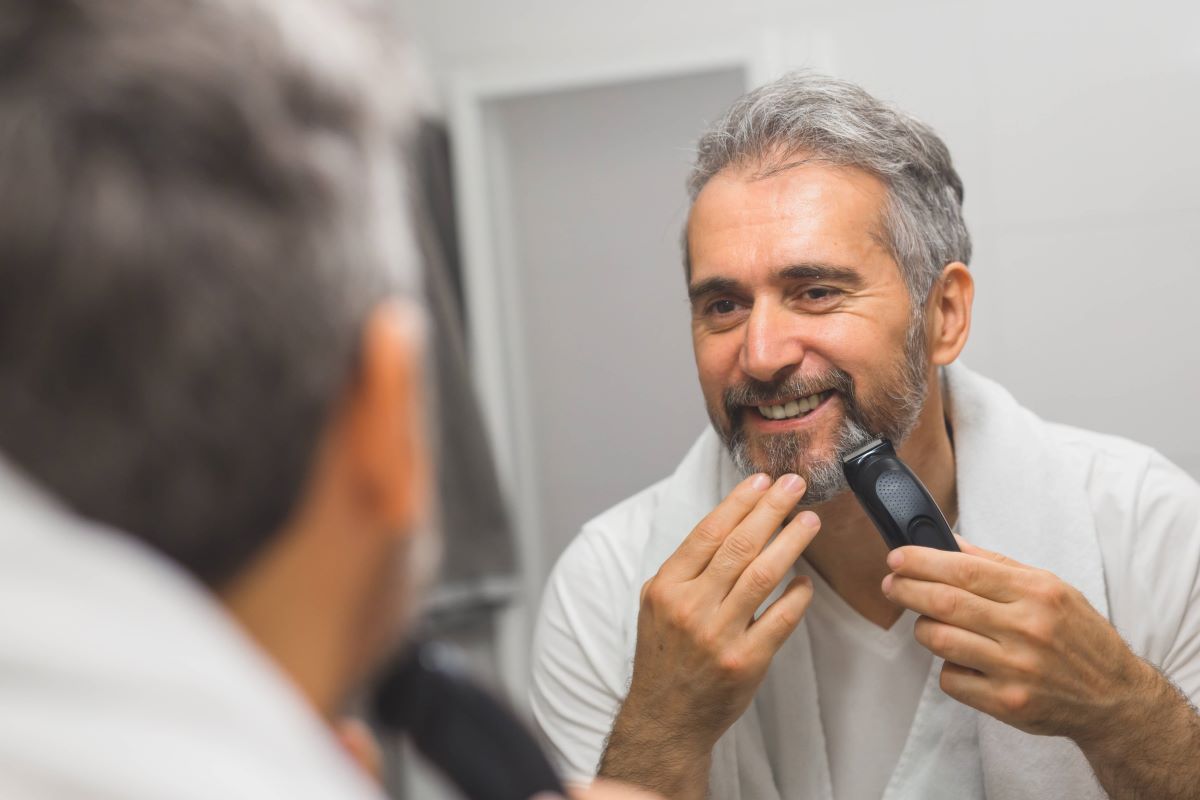 Mann mit grauem Bart trimmt seinen Bart im Spiegel 